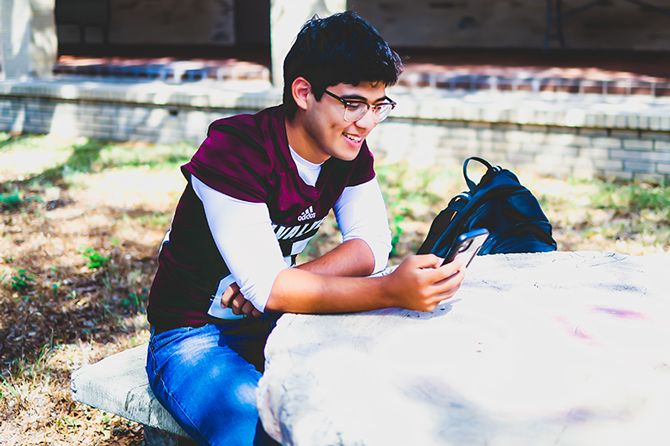 High school student sitting at a bench on campus while looking at their cell phone.