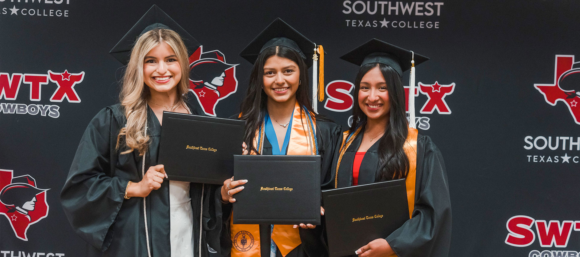 Three graduates pose in front of an SWTX step and repeat holding their diplomas while smiling at the camera.